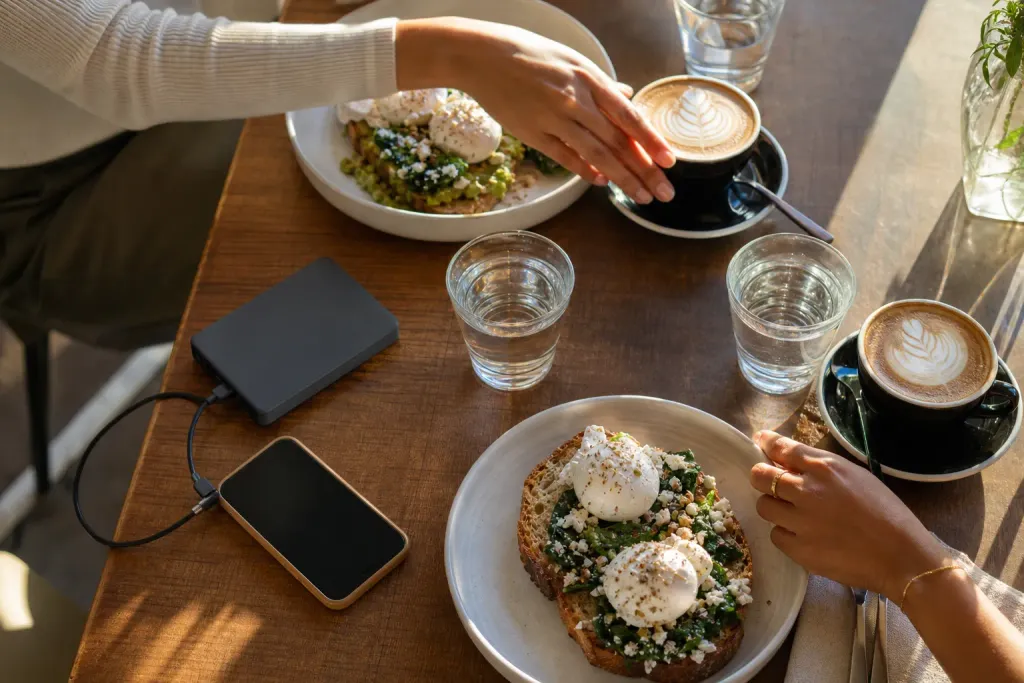 A sunny brunch table with poached eggs, lattes and a phone charging from a MaxCharge power bank